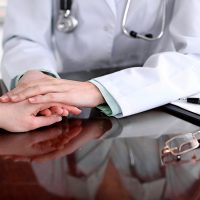 Friendly female doctor hands holding patient hand sitting at the desk for encouragement, empathy, cheering and support while medical examination. Bad news lessening, compassion, trust and ethics concept